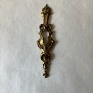 May include: A gold-toned metal keyhole cover with an ornate design. The cover features a decorative top with a pointed finial and a keyhole in the center.