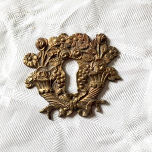 Op de afbeelding: Omsierd messing sleutelgatdeksel met een bloemenontwerp met rozen, druiven en bladeren. Het sleutelgat is omgeven door een decoratieve opstelling van bloemen en bladeren, met een strik aan de onderkant. Het metaal heeft een vintage, antieke uitstraling.
