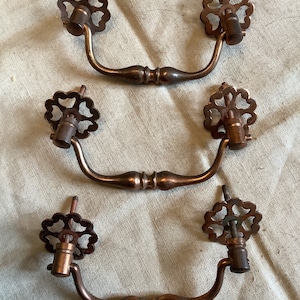 May include: Three antique-style cabinet pulls with a decorative, ornate, clover-shaped design. The pulls are made of a copper-toned metal and have a curved, arched shape.