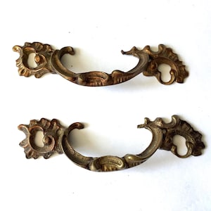 May include: Two antique brass drawer pulls with ornate, scrollwork designs. The pulls have a curved handle and decorative end pieces. The brass has a warm, aged patina, suggesting vintage or vintage-inspired furniture hardware.