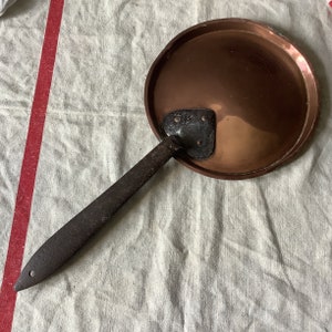 May include: A vintage copper pan with a long, black iron handle. The pan has a rounded bottom and a slightly flared rim.