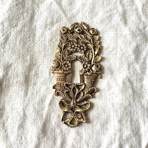 Könnte beinhalten: Verziertes dekoratives Metallstück in Messingoptik mit floralem Design. Das Stück hat eine Schlüssellochform mit Rosen, Blättern und einer Schleife. Das Design ist symmetrisch und detailliert, mit Vintage-Ästhetik.