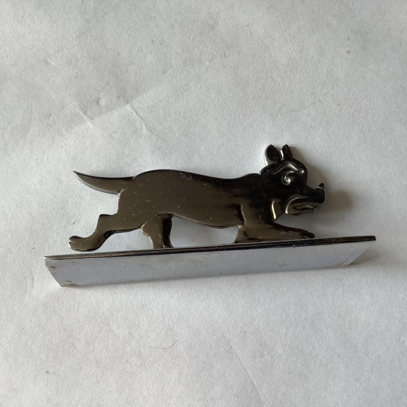 Pode incluir: Uma figura de c&atilde;o cromada, retratada em pose de corrida, montada numa base retangular. As caracter&iacute;sticas do c&atilde;o s&atilde;o detalhadas, com orelhas, focinho e patas vis&iacute;veis. A pe&ccedil;a tem uma superf&iacute;cie reflexiva.