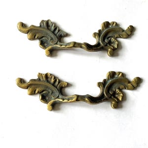 May include: Two matching brass drawer pulls featuring an ornate, scrolling leaf design with a weathered, antique finish. Each handle has a central grip area and two mounting holes positioned within the decorative, wing-like side elements.