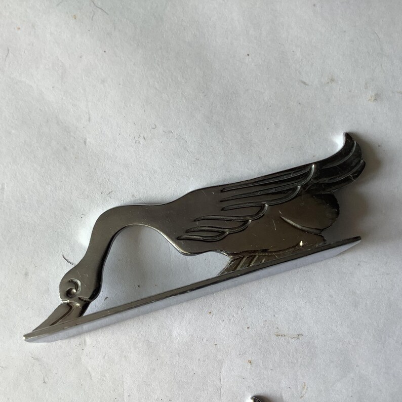 Pode incluir: Um emblema de metal cromado em forma de ave estilizada, possivelmente um ganso ou um cisne, com asas abertas e pesco&ccedil;o curvo. A ave est&aacute; colocada sobre uma base curva.