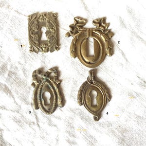 以下が含まれることがあります： 装飾的なデザインが施されたアンティークな真鍮製の鍵穴カバーが4つ。それぞれのカバーは、花柄、リボン、リースなど、異なるデザインが施されています。