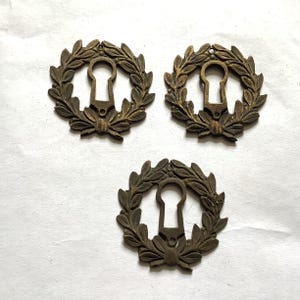 May include: Three antique bronze keyhole covers. Each cover features a keyhole shape surrounded by a laurel wreath design. The keyhole covers are made of metal and have a vintage, aged appearance. The keyhole covers are likely for furniture or decorative purposes.
