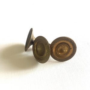 Può includere: Tre antichi candelabri in ottone con dettagli ornati e strutturati. I candelabri hanno una base circolare e una coppa centrale per la candela. L'ottone presenta una calda patina invecchiata, che suggerisce che siano vintage o antichi.