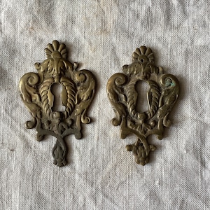 Può includere: Due copri-chiavi in ​​ottone decorati con motivi intricati. Ogni copertura presenta un buco della serratura al centro e un motivo floreale decorativo.
