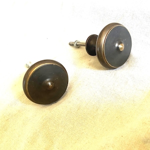 Peut inclure: Deux boutons de meuble anciens en laiton. Chaque bouton a un centre sombre et un bord en laiton. Les boutons sont ronds avec une petite protubérance centrée. Un bouton a un boulon et un écrou attachés pour l'installation. Les boutons sont sur une surface claire.
