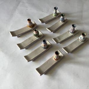 May include: A collection of small, decorative ceramic vases with colorful patterns, displayed on off-white rectangular trays. The vases feature various designs, including floral and geometric motifs. The trays have a slightly curved shape.