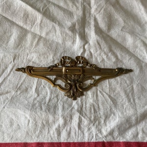 May include: A gold-toned metal decorative trim with a ribbon design and a rectangular cutout in the center. The trim has a detailed, ornate design.