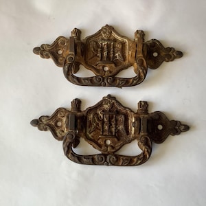 Op de afbeelding: Twee antieke bronzen ladehandgrepen. Elke handgreep heeft een sierlijk ontwerp met een centraal embleem, een gebogen handvat en decoratieve versieringen. De handgrepen zijn verouderd met een verweerde uitstraling, wat wijst op een vintage of gerecyclede oorsprong.