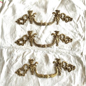 May include: Three ornate gold-toned drawer pulls with a decorative design featuring a face and intricate scrollwork. The pulls are arranged in a row on a white fabric background.