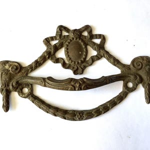 May include: Antique brass drawer pull with ornate details. The pull features a central oval design with a bow above and curved handle. The ends have ram's head designs. A vintage hardware piece for furniture restoration.