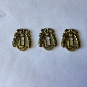 Puede incluir: Tres escudos de cerradura idénticos de latón, cada uno con una abertura vertical central rodeada por un diseño de corona decorativa con un detalle de lazo en la parte superior y un pequeño orificio circular para tornillo en la base.