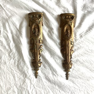 May include: Two ornate gold-colored metal wall sconces with intricate floral and geometric designs. The sconces have a vintage aesthetic and would add a touch of elegance to any room.