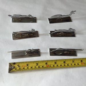 Pode incluir: Uma cole&ccedil;&atilde;o de seis figuras de animais em metal prateado, incluindo um c&atilde;o, um p&aacute;ssaro e um coelho, cada uma montada numa base retangular. Uma fita m&eacute;trica amarela &eacute; colocada perto das figuras, mostrando cent&iacute;metros.