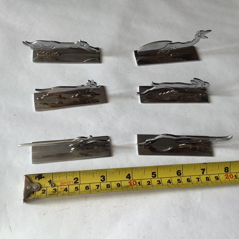 Pode incluir: Uma cole&ccedil;&atilde;o de seis figuras de animais em metal prateado, incluindo um c&atilde;o, um p&aacute;ssaro e um coelho, cada uma montada numa base retangular. Uma fita m&eacute;trica amarela &eacute; colocada perto das figuras, mostrando cent&iacute;metros.