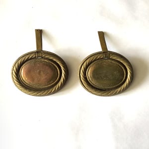 May include: Two antique brass drawer pulls. Each pull has an oval center with a decorative design, surrounded by a rope-like border. A rectangular brass piece extends from the top of each pull.