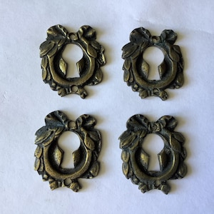 May include: Four antique brass keyhole escutcheons featuring a decorative wreath design with a bow at the top. Each metal plate has a central keyhole opening and two small circular holes for mounting, displaying a dark, weathered patina finish.