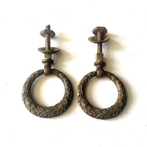 May include: Two antique bronze drawer pulls with ring handles. Each pull features a decorative, textured ring attached to a threaded post with a nut and washer. The pulls have a vintage, aged appearance.