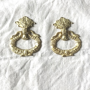 May include: Two ornate, gold-colored drawer pulls. Each pull features a decorative ring with floral and scroll details, connected to a central design with a fan-like top. The pulls are likely for furniture restoration or decorative purposes.