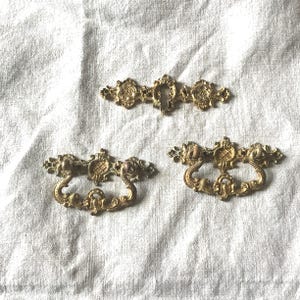 Könnte beinhalten: Drei antike Schubladengriffe aus Messing und eine Schlüssellochblende. Die Schubladengriffe haben verzierte Scroll- und Blumendesigns. Die Schlüssellochblende hat ein passendes Design. Die Hardware ist vor einem weißen, strukturierten Hintergrund platziert.