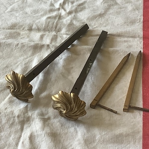 Può includere: Una collezione di ferramenta antica per aste per tende. Due aste hanno terminali decorativi a forma di conchiglia color oro e aste in metallo marrone scuro. Due aste aggiuntive sono semplici, con estremità a punta e finitura dorata. Gli articoli sono disposti su un panno bianco.