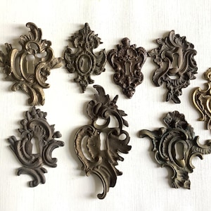 Può includere: Una collezione di otto copri-serratura antichi in ottone con disegni floreali ornati. I copri-serratura variano di colore dal bronzo scuro all'oro, mostrando dettagli intricati e aperture per la serratura. Questi pezzi decorativi sono per il restauro di mobili.