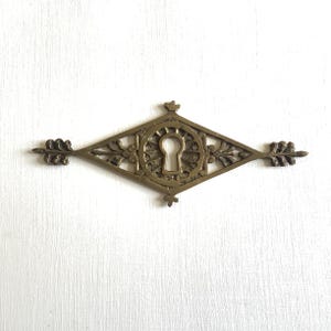 May include: A vintage brass keyhole cover with an ornate diamond-shaped design. The cover features a keyhole in the center, surrounded by floral and leaf details. The ends of the cover have decorative leaf accents. The piece has a warm, antique finish.