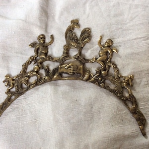 May include: A gold-toned metal decorative piece featuring a rooster, two cherubs, and a hand shake design. The piece is intricately detailed and has a curved shape.