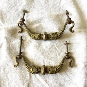 Può includere: Due maniglie antiche per cassetti in ottone con dettagli ornati. Ogni maniglia presenta un'impugnatura curva con abbellimenti decorativi e un punto di montaggio centrale. Le maniglie sono accompagnate dalle loro viti originali.
