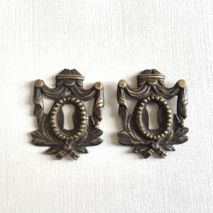 Pode incluir: Duas capas de buraco de fechadura antigas de latão com detalhes ornamentados. Cada capa apresenta um buraco de fechadura rodeado por uma moldura ovalada com contas, com fitas e folhas decorativas. As capas têm uma aparência vintage.