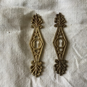 Peut inclure: Deux cache-trous de serrure anciens en laiton doré, ornés de motifs symétriques. Chaque cache-trou présente un centre en forme de losange avec un trou de serrure, entouré de volutes et d'éléments floraux. Couleur or vieilli.
