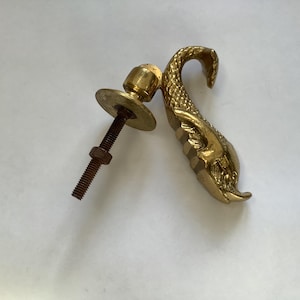 May include: A brass door knocker depicting a mythical sea creature. The knocker is attached to a bolt and washer. The brass has a golden hue. The door knocker is likely for a front door and is a decorative item. The measurements are in centimeters.