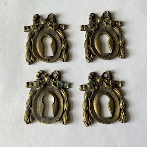 May include: Four antique brass keyhole escutcheons arranged in a grid. Each piece features an oval center opening, a decorative bow at the top, and laurel leaf detailing along the sides. The metal has a tarnished, aged finish with visible patina.