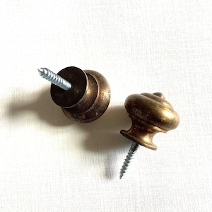Op de afbeelding: Twee antieke messing kastknoppen met bevestigde schroeven. Eén knop is bevestigd aan een ronde basis, terwijl de andere een decoratief, gedraaid ontwerp heeft. De knoppen hebben een warme, verouderde goudkleur.