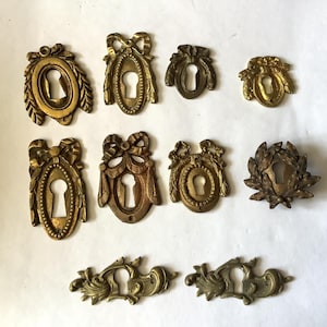 Könnte beinhalten: Eine Sammlung antiker Messing-Schlüssellochabdeckungen in verschiedenen verzierten Designs. Die Abdeckungen weisen dekorative Elemente wie Schleifen, ovale Rahmen und florale Motive auf. Das Messing hat eine warme, gealterte Patina.