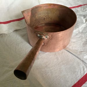 Peut inclure: Une casserole en cuivre vintage avec une longue poignée. La casserole a une patine usée et vieillie et est légèrement bosselée.