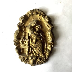 May include: A gold-colored metal wall plaque with an ornate design featuring a relief sculpture of the Virgin Mary holding a baby Jesus.