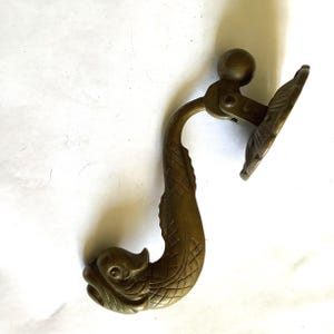 May include: A brass-colored metal drawer pull featuring a curved dolphin-shaped handle with etched scale details. The handle is attached to a circular, scalloped mounting plate by a hinged joint, allowing the dolphin body to swing freely.