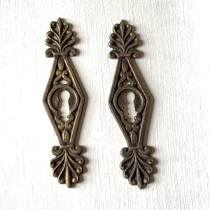 Puede incluir: Dos cubiertas de cerradura antiguas de bronce con diseños florales y geométricos ornamentados. Cada cubierta presenta una forma de diamante con un ojo de cerradura en el centro y adornos decorativos en la parte superior e inferior. Fondo blanco.