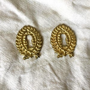 Könnte beinhalten: Zwei antike Schlüssellochabdeckungen aus Messing. Jede Abdeckung ist oval, von einem Lorbeerkranz-Design umgeben und hat ein Schlüsselloch in der Mitte. Das Messing hat einen warmen, goldenen Farbton, der auf Alter hindeutet. Ideal für die Restaurierung antiker Möbel.