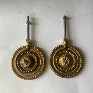 Peut inclure: Deux poignées de tiroir antiques en laiton. Chaque poignée circulaire présente un motif floral central, entouré de bordures perlées et en forme de corde. Une tige filetée s'étend du haut pour le montage. Patine chaude et vieillie.