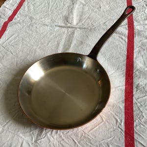 以下が含まれることがあります： 長いダークブラウンのハンドルが付いた銅製のフライパン。フライパンは光沢のある滑らかな表面をしています。