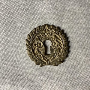 May include: A gold-toned metal keyhole cover with a decorative floral design.