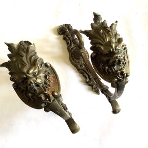 Könnte beinhalten: Zwei antike Vorhanghalter aus Messing mit einem detaillierten Löwenkopfmotiv und verzierten Blattakzenten. Jedes Metallbeschlagteil hat ein geschwungenes Hakendesign und eine verwitterte Patina, die komplizierte Reliefarbeiten zeigt.