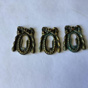 May include: Three antique brass keyhole escutcheons featuring a decorative bow at the top and a beaded oval frame surrounding the central keyhole opening. Each piece displays a weathered, tarnished metal finish with visible green oxidation.
