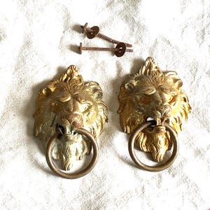 May include: A pair of antique brass lion head door knockers with ring handles. The lion heads feature detailed manes and facial features. Two screws are included. The knockers have a vintage, aged appearance, with a golden hue.